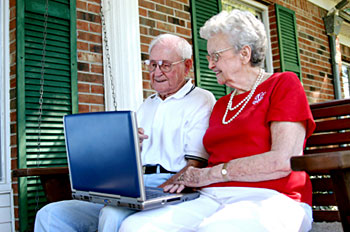 An elderly couple sitting in a swing on their front porch, both looking at a laptop computer