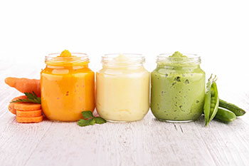 three clear glass bottles of baby food including pureed carrots and peas