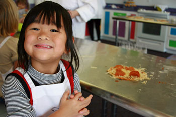 a child with long dark hair stands proudly next to the pizza she made in cooking class