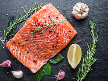 a fresh filet of salmon on a cutting board accompanied by cloves of garlic, a wedge of lemon, and rosemary