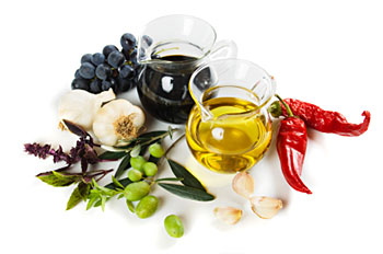 a carafe of balsamic vinegar and a carafe of olive oil, flanked by garlic, fresh green olives, and red peppers