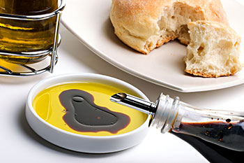 Balsamic vinegar being poured into a dish already containing olive oil. Bread is nearby for dipping.