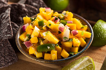 a bowl of mango salsa with blue corn chips and slices of lime on a wooden table