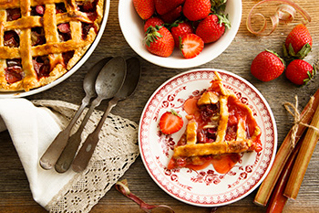 a strawberry rhubarb pie with a slice cut out of it flanked by fresh strawberries and rhubarb stalks