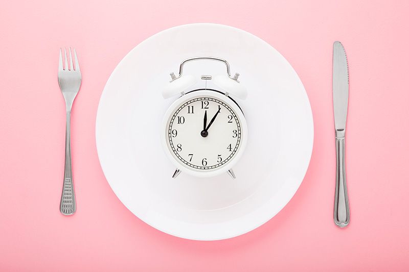 an alarm-style clock set to 12:06 on a plate flanked by a knife and fork
