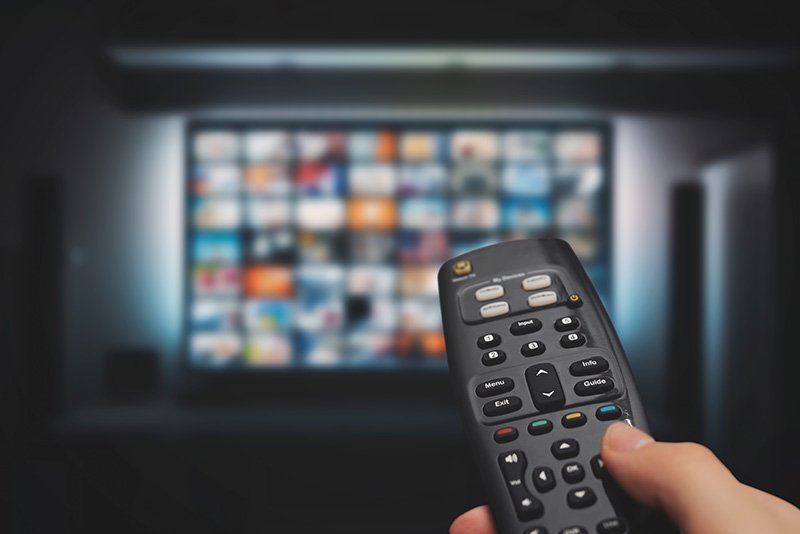 a person's hand holding a remote control, which is pointed at a large-screen television.